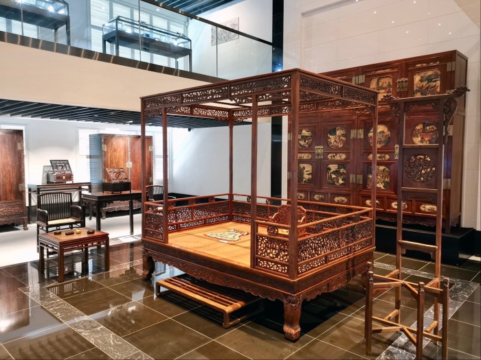 Antique wooden furniture including lounge chairs, a canopy bed and a cabinet, are on display in a museum room