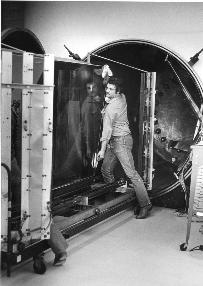 Black and white photo of a man heaving a pane of glass into a large machine