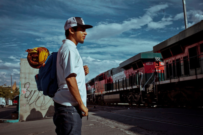 A man in a hat and with  a bag and blanket on his back watches a heavy-goods train passing