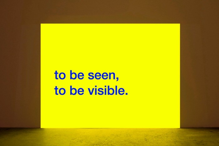 A rectangular yellow projection on a wall with the words in blue ‘to be seen, to be visible’