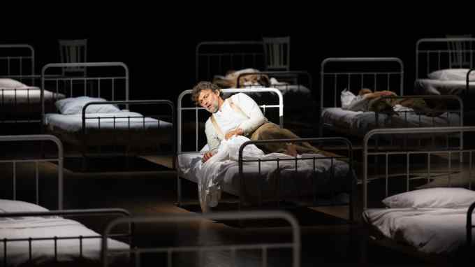 A man lies semi-prone on a metal hospital bed amid rows of identical beds