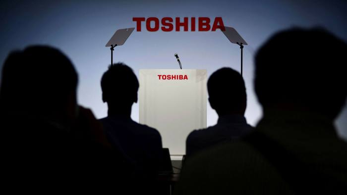 An audience of four in silhouette look at a stage with the Toshiba logo, a lectern and two teleprompters