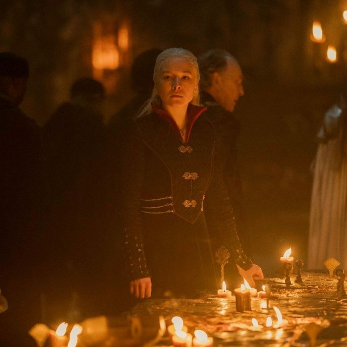 A woman with white-blonde hair stands looking sombre in a dark room next to a table where numerous candles cast a glow