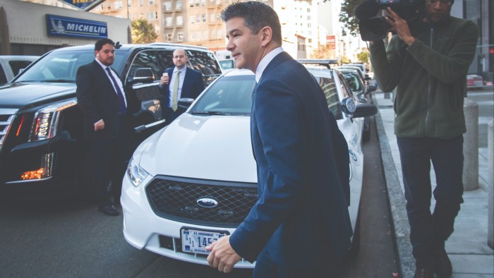 Travis Kalanick, co-founder and former chief executive officer of Uber Technologies Inc., exits a building. In the background are some cars, some onlookers and a cameraman 