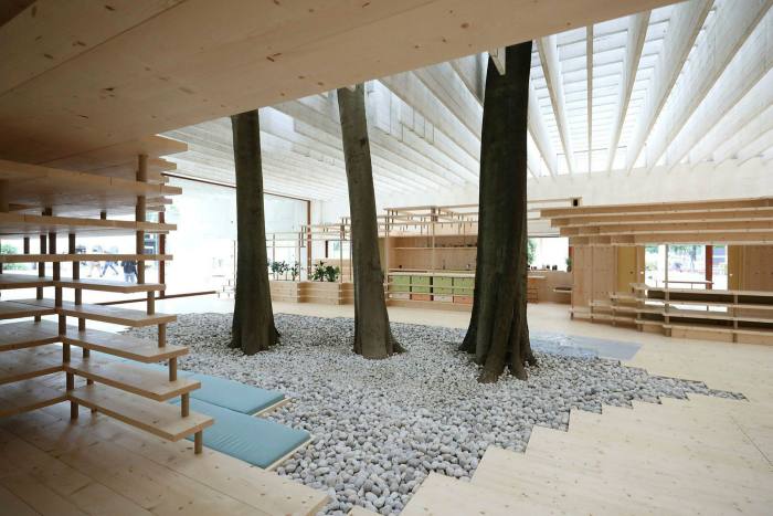 The interior of a building with a slatted roof and wooden fixtures. In the centre are three tree trunks surrounded by a grey pebble floor