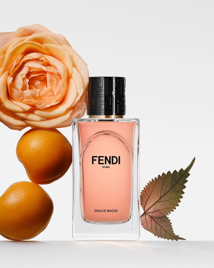 An orange-coloured rectangular bottle with black shiny lid stands in the centre with an orange rose and two oranges on one side of it and two leaves on the other