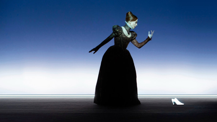 A woman in a long Elizabethan-style dress faces sideways on a bare stage; in front of her is a white shoe