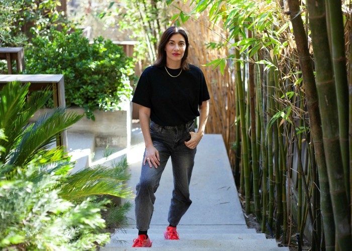 A severe-looking woman in a black T-shirt standing on concrete steps in sunlight filtered through a bamboo wall