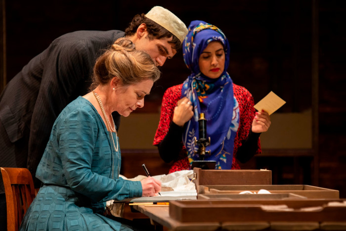 A middle-aged woman in a turquoise dress sits writing at a table while a man dressed as an Arab servant looks over her shoulder and a a woman with a hijab over he head sits in the background 