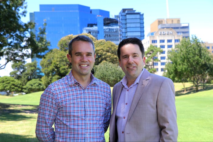 Portrait of two men standing side-by-side in a park with office blocks in the distance