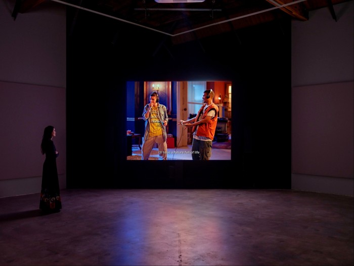 A young woman standing on the left of an audiovisual installation in a darkly lit room watches two young rappers sing
