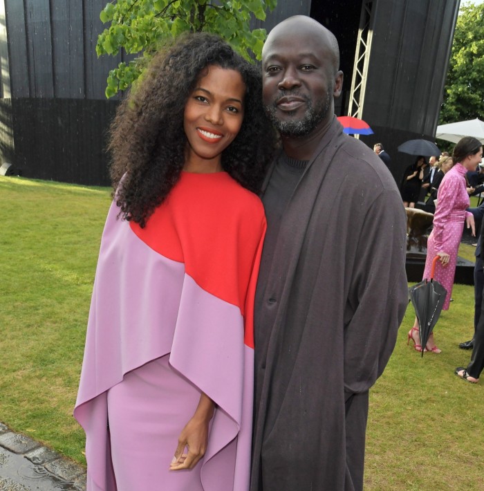 Ashley Shaw Scott, wearing a flowing pink and red dress, stands next to David Adjaye, who is wearing a dark suit 