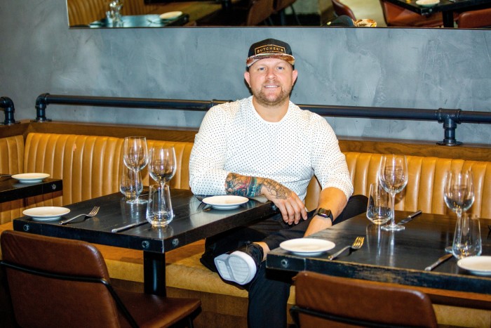 Stubborn Seed chef/owner Jeremy Ford sitting on a coffee-coloured banquette in his restaurant. He is wearing a white dot-pattern jersey, black trousers, trainers and baseball cap, and has a goatee beard