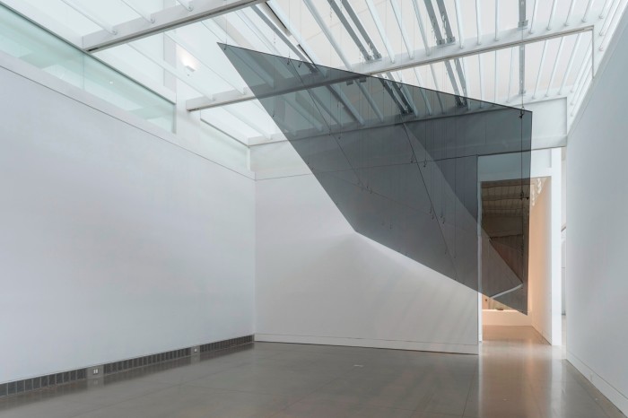 A rectangular, thin and black glass sculpture hangs obliquely from the ceiling of a gallery.