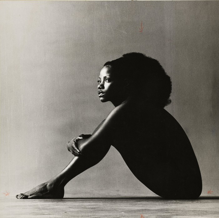 A nude woman sits on the floor, silhouetted against a grey background, so that her body is almost entirely black