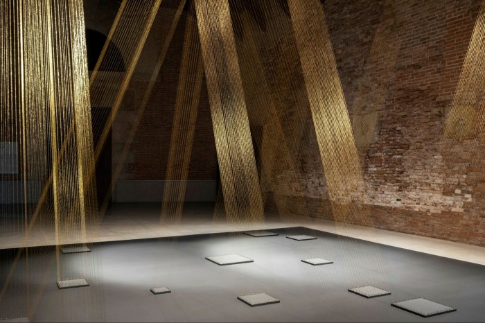 Golden threads stretch from ceiling to floor inside a gallery like shafts of sunlight