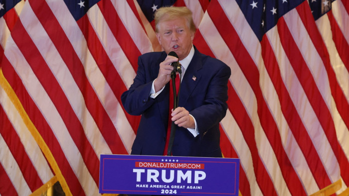 A man in suit stands at a podium with ‘Trump’ written on it and with American flags in the background