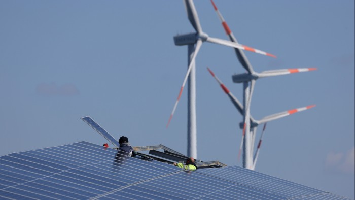 Workers install solar panels as wind turbines spin behind them