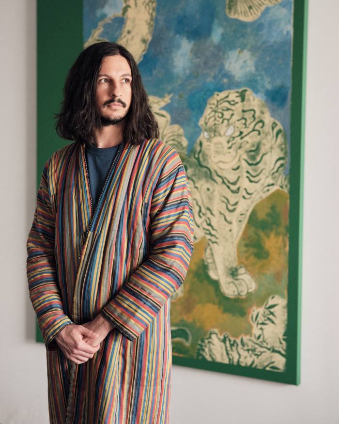 Kour Pour wears a coat he made, in front of a work-in-progress