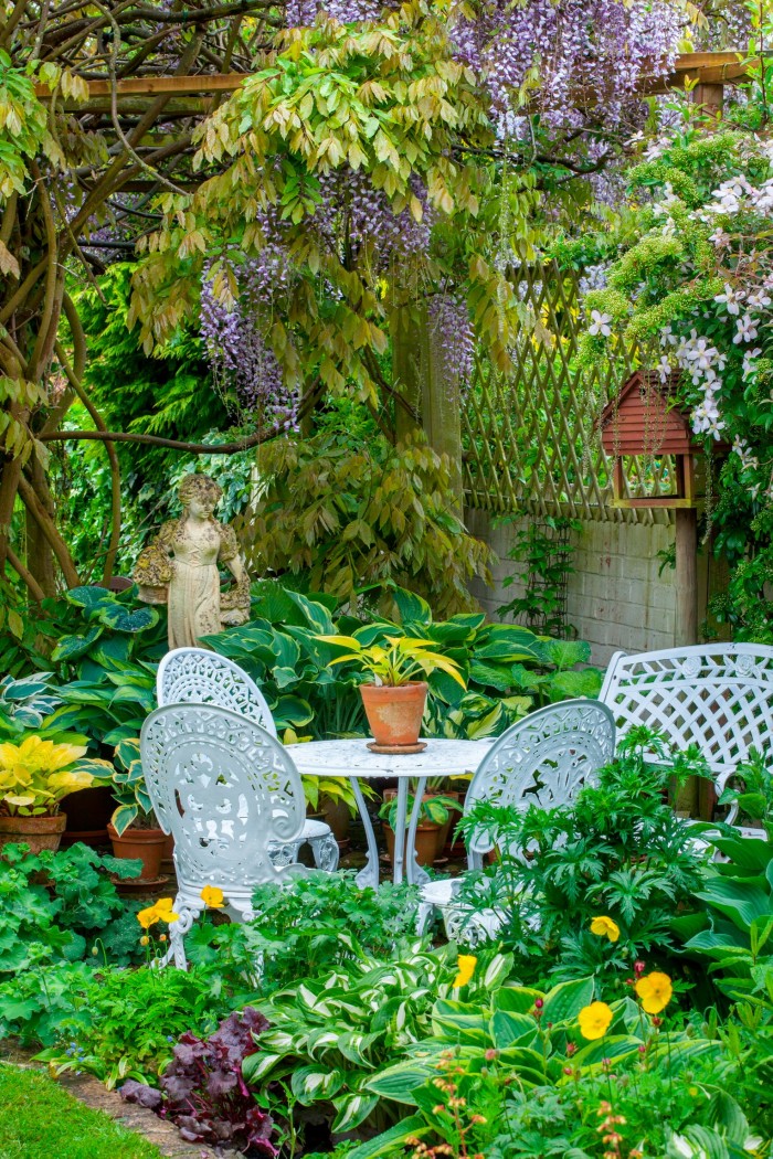 White metal table and chairs on patio under wisteria, hostas in containers
