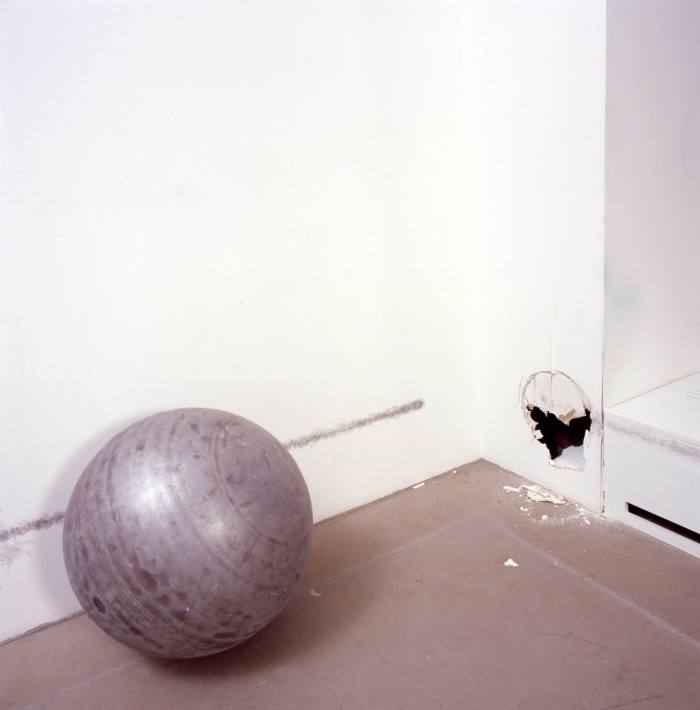 A small steel ball sits on a gallery floor next to a white wall with a hole in it