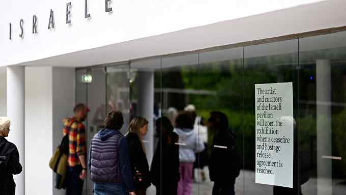 People in coats stand outside a modern building with a white facade and the word ‘Israel’ above their heads. In the shop window, a notice reads: ‘The artists and curators of the Israeli pavilion will open the exhibition when a ceasefire and hostage release agreement is reached’
