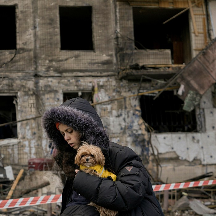 A woman in a hooded black parka carries a small dog as she walks past the wreck of a building destroyed by shelling