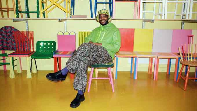 A man in a bright green jacket and grey trousers sits among colourful chairs