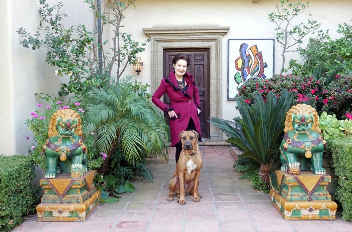 A woman in a smart cerise jacket standing behind a dog and in front of a large house