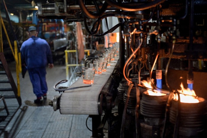 A factory engineer walks past a conveyer belt of glass on a furnace