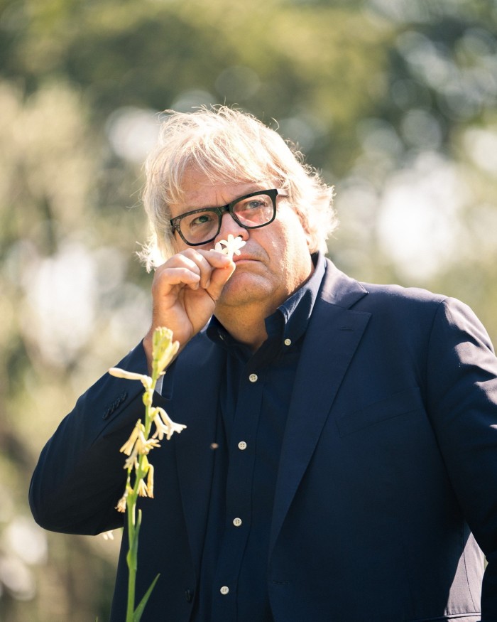 A portrait of a bespectacled middle-aged man with blond hair, dressed in a dark suit and navy shirt, smelling a tiny white flower against a blurred natural backdrop.