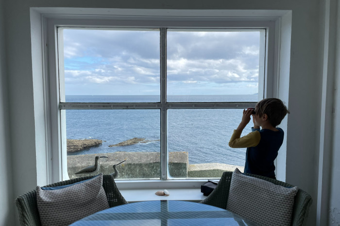 A boy looks out of a wide window at a view of the sea with the aid of binoculars 