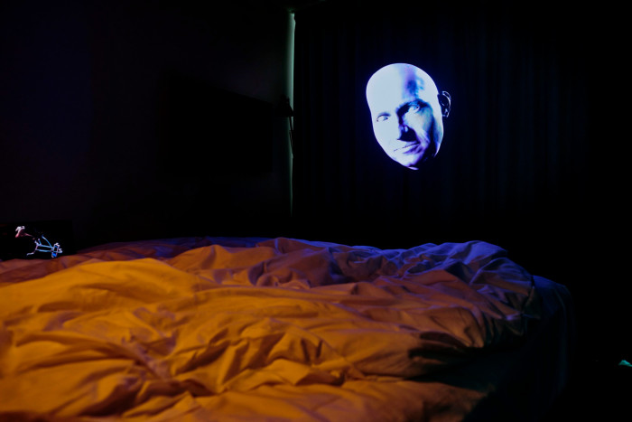 In a scarcely lit room, the light from a projection showing a bodyless white head turns the bedsheets in the foreground in a warm swathe of orange and purple