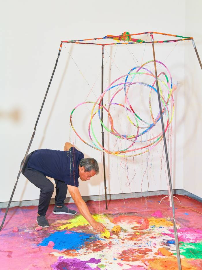 The artist crouches underneath a colourful hanging sculpture supported by a frame. The floor is splashed with paint. 