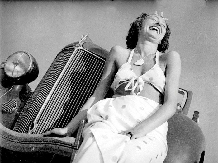 A woman in a bikini top sitting on the front of a car laughing
