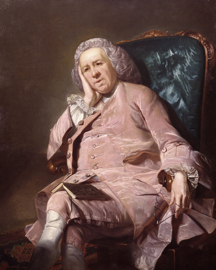 A weary looking man in a pink suit sits back in an armchair, his head resting on one hand