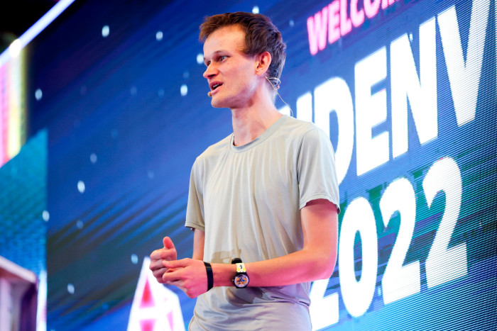 Vitalik Buterin wears a white T-shirt and addresses delegates from the stage at a blockchain event