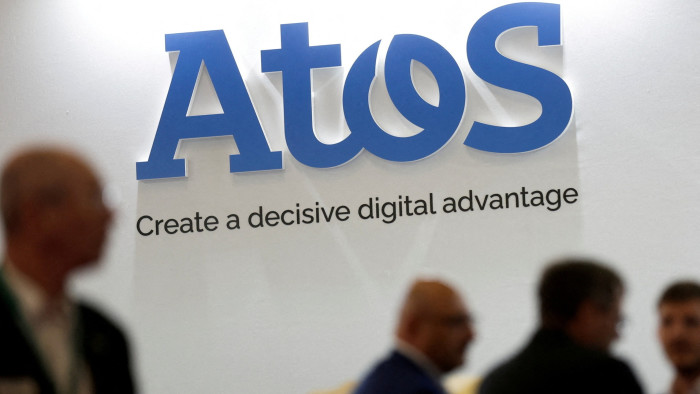 People pass in front of the Atos logo on a wall. A slogan underneath reads: ‘Create a decisive digital advantage’