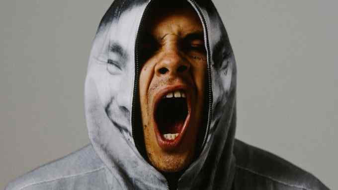 A portrait photo of Slowthai with his mouth open as if shouting and face emerging through an unzipped hood printed with his smiling face