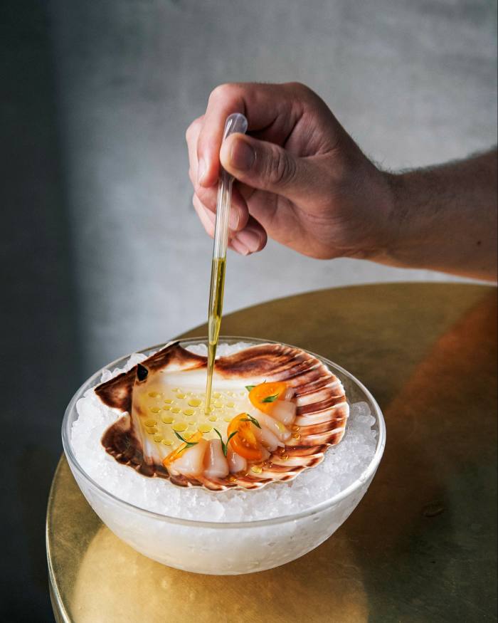 A hand holding a pipette of olive oil over a dish on a bowl of ice of Orkney scallop with tomato essence