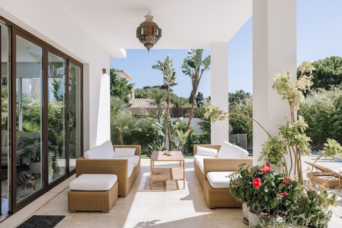the covered terrace of a whitewashed villa surrounded by sunshine, plants and palm trees