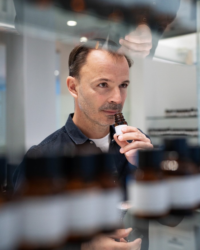 A man in a denim shirt is intently smelling a dark bottle, partially obscured by rows of bottles on glass shelves