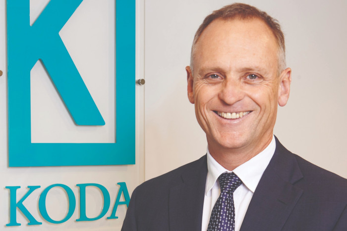 Smiling businessman in a formal navy suit, white shirt, and patterned blue tie, standing beside a company logo sign that reads ‘KODA’
