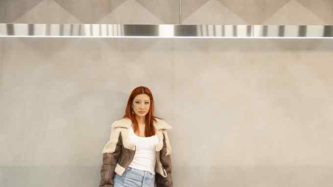 An Asian woman with auburn hair and wearing a furry flight jacket stands against a beige wall