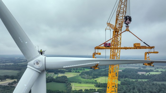 A large  mobile crane lifts a rotor blade to the nacelle of a wind turbine