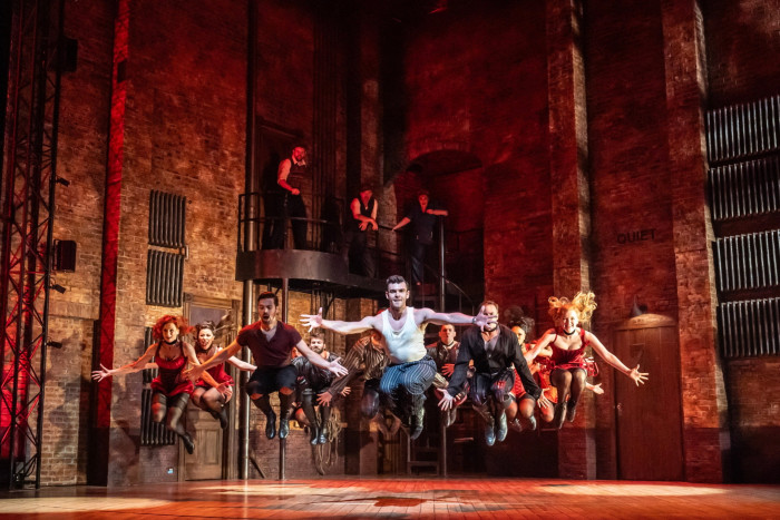 In a high, brick-walled space, a group of male and female dancers leap athletically and grin; behind them three people look down on them from a balcony