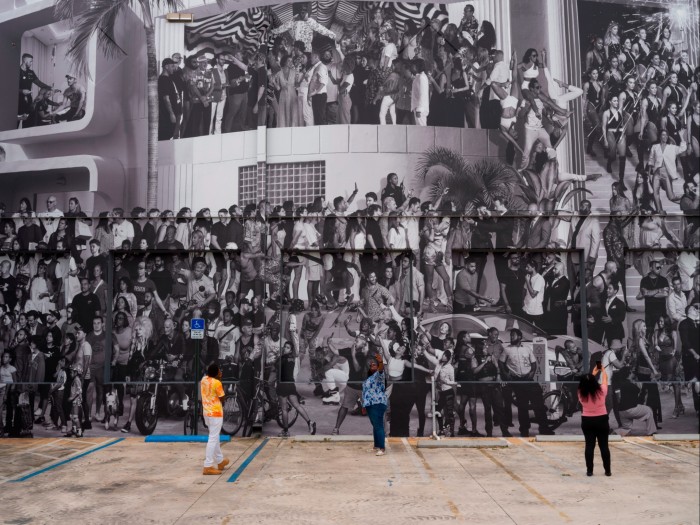People take selfies in front of a large black and white photographic mural crowded with figures