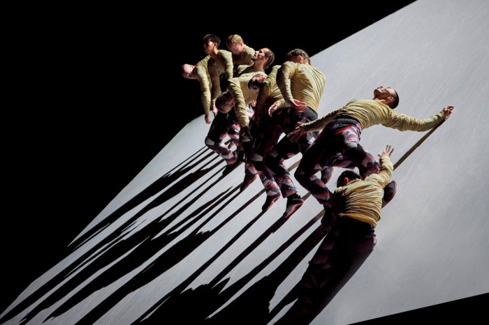 A group of dancers stand in various positions on a large steeply tilting platform; the lighting casts long downward shadows