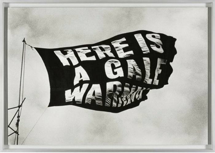 A black and white photo of a black flag waving. It has white writing reading: here is a gale warning
