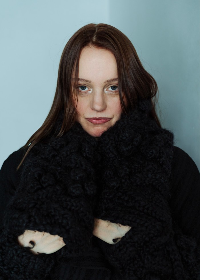 A young woman wearing a black top and a black knitted scarf stares into the camera